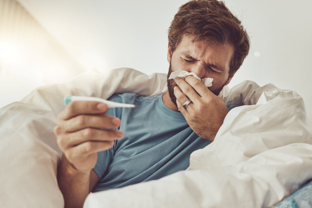 Mann mit Fieberthermometer in der Hand liegt im Bett und putzt sich die Nase