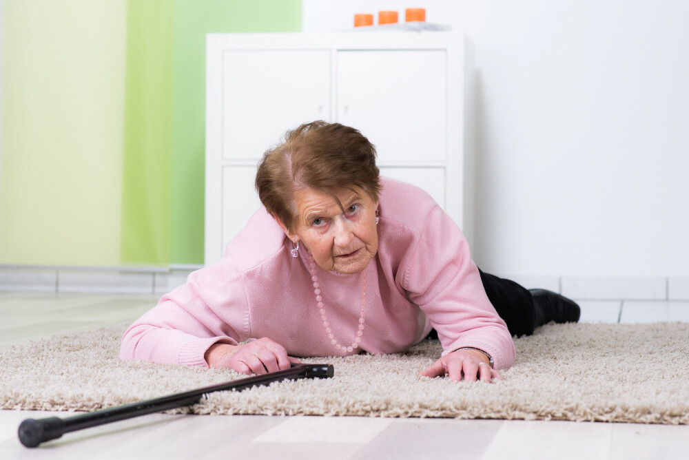 Seniorin in rosfarbenem Pullover ist auf Teppich gestürzt. Im Vordergrund liegt ein Gehstock