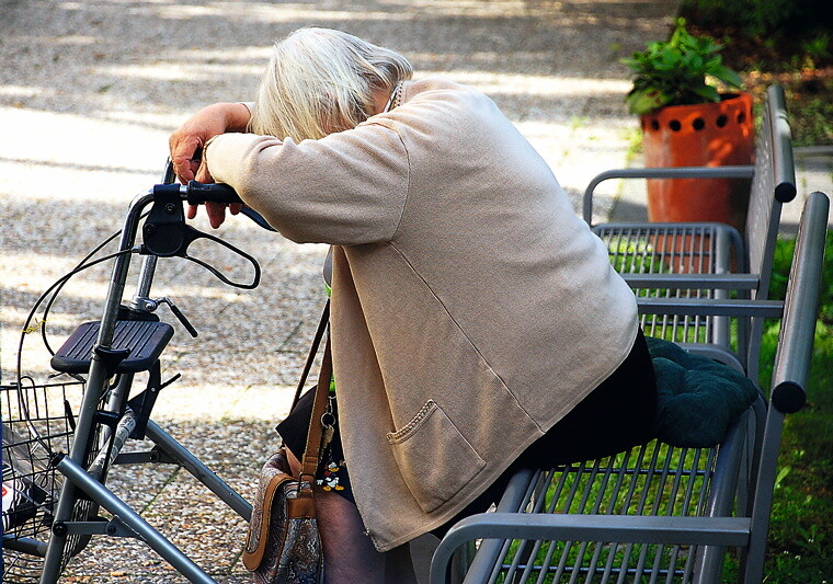 Ältere Frau mit Rollator sitzt auf einer Parkbank.