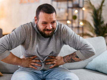 Mann mit schmerzhaft verzerrtem Gesicht sitzt auf dem Sofa und hält sich den Bauch